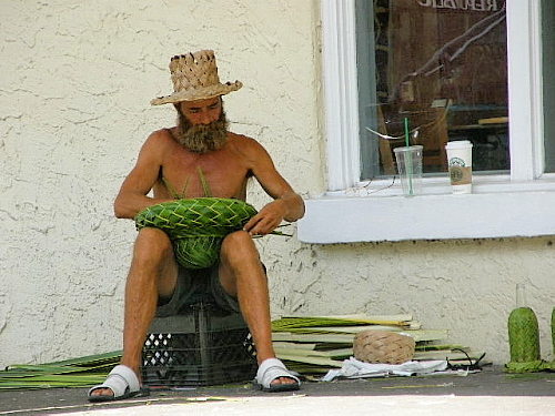 street merchant weaving baskets from palm leaves sitting shirtless on a wooden box a straw hat shading his face as he is intent on the task